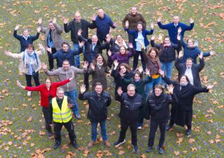 Eine Gruppe von rund 30 Personen steht im Freien auf einem mit Laub bedeckten Boden. Alle Personen heben die Hände und lächeln in die Kamera. Einige tragen verschiedene Kleidungsstile, darunter formelle und lässige Kleidung.