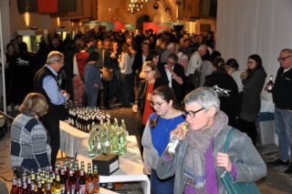 Eine große Menschenmenge in einem Veranstaltungsraum, die sich um Tische mit Getränken versammelt hat. Im Vordergrund stehen mehrere Flaschen mit Limonade und Wasser, während im Hintergrund Gäste miteinander sprechen und anstoßen.