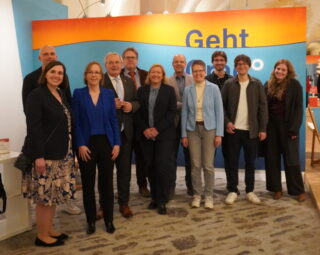 Eine Gruppe von neun Personen steht in einem Ausstellungsraum mit blauer Wand. Im Hintergrund ist ein Banner mit der Aufschrift "Geht" sichtbar. Alle Anwesenden lächeln und tragen formelle Kleidung. Der Raum hat hohe Decken und ein rustikales Ambiente.