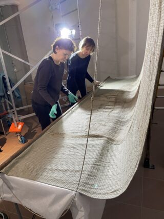 Zwei Frauen arbeiten zusammen, um ein großes, faltiges Gewebe auf einem Tisch zu glätten. Die Frauen tragen grüne Handschuhe und sind in einem Raum mit künstlicher Beleuchtung und geräumigem Hintergrund, der ein Gestell zeigt.