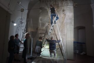 Eine Person steht auf einer Leiter und arbeitet an einer großen Wandmalerei in einem Ausstellungsraum. Um sie herum beobachten mehrere andere Personen das Geschehen vor der Wand, die mit historischen Symbolen und Texten verziert ist.