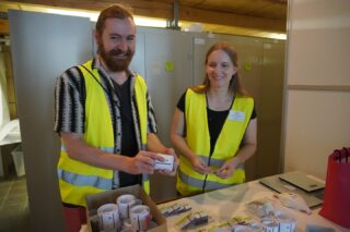 Ein Mann und eine Frau in gelben Warnwesten stehen an einem Tisch mit Rollen, Stiften und kleinen Packungen vor grauen Schränken.