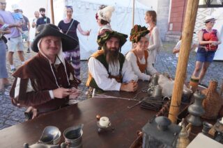 Drei Personen in historischer Kleidung sitzen an einem Holztisch mit Metallgeschirr und Gefäßen, im Hintergrund stehen weitere Besucher auf einem gepflasterten Platz.