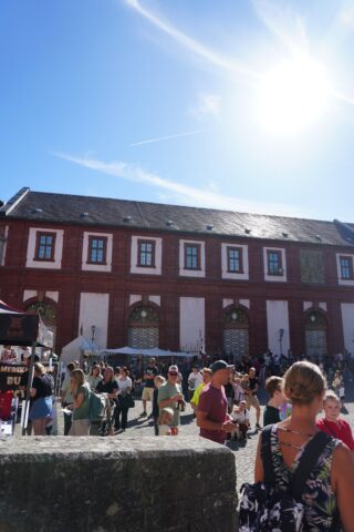 Eine Menschenmenge steht vor einem langen roten Backsteingebäude mit weißen Fenstern, während die Sonne hoch am blauen Himmel scheint.