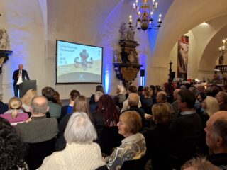 Eine Menschenmenge sitzt in einem Saal, während Benjamin Spies die Veranstaltung eröffnet. Auf dem Bildschirm ist die Ankündigung „Lange Nacht der Wissenschaft“ zu sehen.