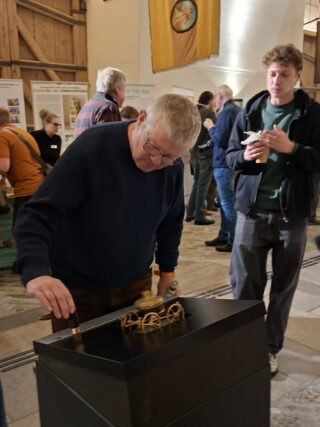 Ein Mann untersucht eine bewegliche Ausstellung, indem er daran arbeitet. Im Hintergrund sind weitere Besucher zu sehen, während Informationen an Wänden hängen. Der Raum ist hell erleuchtet und hat eine rustikale Atmosphäre.