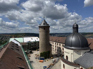 Ein Blick auf eine historische Burg mit einem runden Turm und einer Kuppel, umgeben von Baugerüsten. Wolken sind am Himmel zu sehen, während die Landschaft im Hintergrund sichtbar ist.