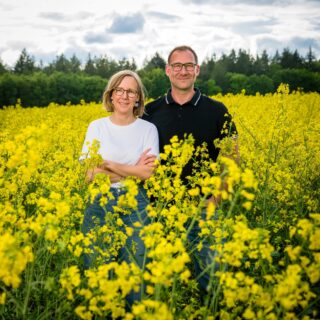 Ein lächelndes Paar steht in einem gelben Rapsfeld vor einer Baumlinie und bewölktem Himmel.