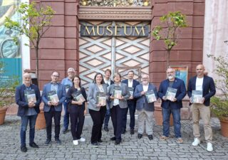 Eine Gruppe von zwölf Personen, die Broschüren in der Hand halten, steht vor einem Museumseingang mit dem Schriftzug "MUSEUM". Die Teilnehmer tragen formelle Kleidung und stehen in einer gepflasterten Fläche mit Topfpflanzen im Vordergrund.