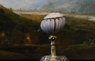 Ein Trinkpokal vor einem Gemälde. Er ist Gold und weiß. Oben ist ein Pferd eingraviert.