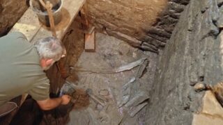 Ein Archäologe untersucht eine Ausgrabungsstelle in einem Schacht. Im Schacht sind Holzreste und Erde sichtbar. Der Boden ist uneben und das Mauerwerk ist gut erhalten. Werkzeuge befinden sich auf einem Tisch am Rand der Ausgrabung. Foto: © PD Dr. Markus Schußmann