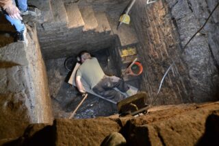 Ein Mann in Arbeitskleidung liegt in einer tiefen Grube und arbeitet mit einem Werkzeug. Die Wände der Grube sind aus Stein, und es gibt einige Werkzeuge und eine orangefarbene Eimer im Hintergrund. Ein weiterer Teil der Stufen ist sichtbar, sowie eine Person, die über die Grube schaut.
