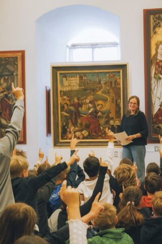 Eine Frau steht vor einer Gruppe von Kindern in einem Ausstellungsraum. Die Kinder heben begeistert die Hände, während hinter ihnen ein großes Gemälde mit religiösen Motiven an der Wand hängt. Natürliches Licht fällt durch ein Fenster.