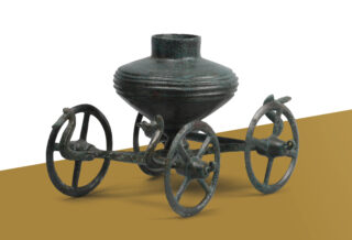 Ein antikes bronzenes Gefäß auf einem vierrädrigen Gestell. Das Gefäß hat einen runden Körper mit horizontalen Rillen und einen kurzen Hals. Die Räder sind detailreich gestaltet und das Gefäß steht auf einer diagonalen, braunen Fläche.