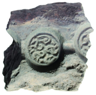 Ein unregelmäßig geformter Steinblock mit zwei kreisförmigen reliefartigen Mustern auf der Vorderseite. Die Muster zeigen geschwungene Linien, die in einem symmetrischen Design angeordnet sind.