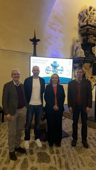 Vier Personen stehen in einem Raum mit steinernen Wänden. Im Hintergrund ist ein Bildschirm mit der Aufschrift "Lange Nacht der Wissenschaft" sichtbar. Die Personen sind formal gekleidet; zwei Männer tragen Blazer, eine Frau in der Mitte trägt eine rote Bluse. Licht strahlt auf die Szene.