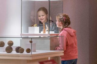 Ein Mädchen schaut fasziniert auf Ausstellungsstücke in einem Museumsdisplay. Im Hintergrund sind weitere Objekte sichtbar. Die Atmosphäre ist hell und einladend.