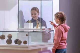 Zwei Mädchen stehen an einem Vitrinenstand in einem Museum. Eines der Mädchen zeigt mit dem Finger auf eine Statue in der Vitrine, während das andere Mädchen sie lächelnd betrachtet. Im Hintergrund sind weitere Objekte in der Vitrine sichtbar.