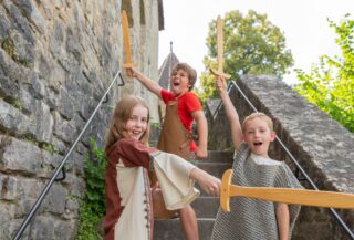 Drei Kinder stehen auf einer Treppe aus Stein und halten Holzschwerter in die Höhe. Das Kind links trägt ein braunes und rotes Gewand, das in der Mitte ein rotes T-Shirt mit einer grünen Weste zeigt, und das Kind rechts trägt ein graues T-Shirt, während die Umgebung von Natur und einem historischen Gebäude geprägt ist.