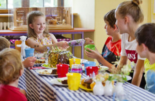 Eine Gruppe von Kindern sitzt an einem Tisch mit bunten Tellern und Tassen, während ein Mädchen mit einem Lächeln einen Kuchen serviert. Der Tisch ist festlich gedeckt und bietet eine Vielzahl von Speisen.