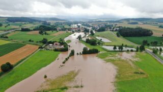 Ein Luftbild einer überfluteten Landschaft, in der Weiden und Felder unter Wasser stehen. Im Hintergrund sind kleine Häuser im Dorf zu sehen, umgeben von Bäumen und hügeliger Wiese. Der Himmel ist bewölkt.