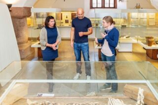 Drei Personen stehen in einem Museum um eine verglaste Ausstellungsfläche, im Hintergrund stehen helle Vitrinen und Fenster.