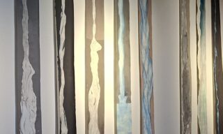 Eine Wand mit mehreren vertikalen Streifen aus gemalten Materialien in verschiedenen Grau- und Blautönen. Die Streifen sind unterschiedlich breit und zeigen fließende Formen, die an Wasser oder Wellen erinnern.
