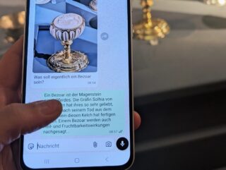 Eine Hand hält ein Smartphone, das eine Nachricht über goldene Objekte anzeigt, während im Hintergrund verschiedene goldene Gefäße und Tassen in einer Vitrine zu sehen sind.