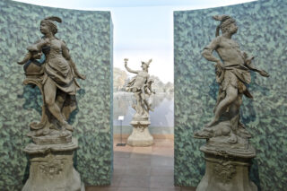 Drei Skulpturen in einem Museum, zwei Skulpturen stehen auf Podesten. Links eine weibliche Figur mit einem Gefäß und rechts eine männliche Figur mit einer Fackel, während in der Mitte eine weitere Skulptur im Hintergrund sichtbar ist.