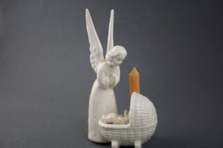 Eine weiße Statue eines Engels mit gefalteten Händen, die über einen Korb mit einem kleinen Kind schaut. Neben dem Korb steht eine orangefarbene Kerze. Der Hintergrund ist dunkelgrau.