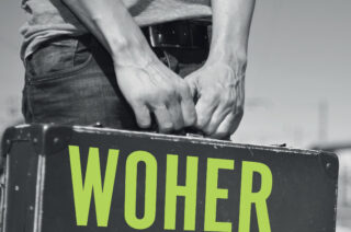 Ein Mann hält einen schwarzen Koffer mit der Aufschrift "WOHER" in großen grünen Buchstaben. Seine Hände sind sichtbar, während er den Koffer umschließt. Der Hintergrund ist unscharf und scheint eine Außenumgebung zu sein.