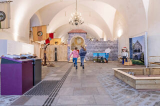 Zwei Kinder laufen in einem Museum, das mit verschiedenen Ausstellungsstücken dekoriert ist. Im Hintergrund sind thematische Wandbilder und ein Bereich mit Holzblöcken zu sehen.