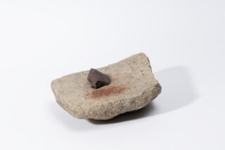 Ein graues, flaches Gestein mit einer kleinen, spitzen, dunklen Mineralienbordüre in der Mitte. Das Gestein hat eine raue Oberfläche und liegt auf einem weißen Hintergrund.