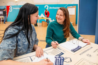 Zwei Frauen sitzen an einem Tisch und lächeln sich an. Die Frau links trägt ein graues Oberteil und hat lange schwarze Haare, während die Frau rechts ein grünes Oberteil trägt und kurze braune Haare hat. Auf dem Tisch liegen mehrere Dokumente und Schreibutensilien.