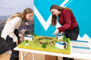 Zwei Frauen betrachten ein Modell einer historischen Anlage mit Holzpalisaden. Das Modell steht auf einem Tisch, umgeben von kleinen Bäumen. Eine Infotafel mit Text ist rechts neben dem Modell platziert. Im Hintergrund ist eine graue Wand mit Landschaftsbildern.