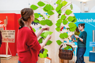 Eine Frau in einem roten Pullover hängt grüne Blätter an einen baumartigen Ausstellungsstand. Im Hintergrund ist eine weitere Person zu sehen, die an einer anderen Station arbeitet. Die Umgebung wirkt hell und einladend.