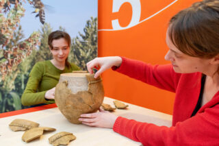 Zwei Frauen arbeiten an einem Tontopf auf einem Tisch. Eine Frau in rotem Pullover setzt vorsichtig ein Stück auf den Topf, während die andere Frau in grünem Oberteil im Hintergrund sitzt und zuschaut. Der Hintergrund ist orange mit einer großen Zahl 5.