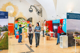 Zwei Frauen gehen durch eine Ausstellung mit farbigen Wänden und Informationsständen. Im Hintergrund arbeiten mehrere Personen an Tischen. Der Raum hat hohe Decken und ist mit einem großen Lichtobjekt dekoriert.