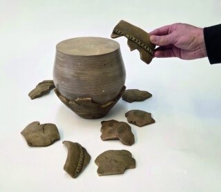 Ein Mensch hält ein Stück eines zerbrochenen Tontopfs und betrachtet den unbeschädigten Topf. Um den Topf herum liegen viele Tonstücke, die von der Zerbrechung stammen.