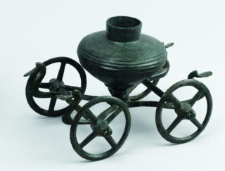 Ein antikes, bronzenes Gefäß auf einem Wagen mit vier Rädern. Das Gefäß hat eine runde Form und einen schmalen Hals. Die Räder sind mit einem Kreuzmuster gestaltet und ermöglichen das Rollen des Wagens.