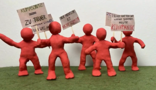 Fünf rote Knetfiguren halten Schilder mit Aufschriften zu Klimaschutz und Klimawandel. Die Figuren stehen auf einer grünen Fläche vor einer weißen Wand. Die Schilder tragen Texte wie "Zu teuer" und "Der Mensch ist nicht schuld".