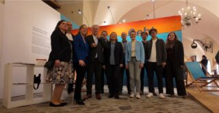 Eine Gruppe von zwölf Personen steht auf einer Galerie, umgeben von hellen Wänden und einem großen Banner mit dem Wort "lebt". Die Personen sind in formeller Kleidung und posieren für ein gemeinsames Foto. Im Hintergrund sind Kunstwerke und eine große Lampe sichtbar.