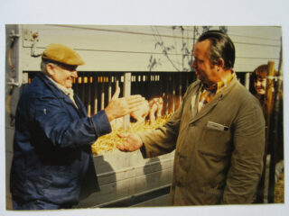 Zwei Männer schütteln sich die Hände vor einem Transporter, aus dem vage Silhouetten von Tieren sichtbar sind. Der Mann links trägt eine Mütze und einen blauen Overall, der Mann rechts hat eine Jacke und eine Krawatte an.