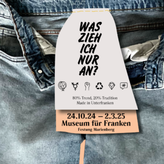 Nahaufnahme von einer Jeans mit einem Etikett, das die Frage "Was zieh ich nur an?" stellt. Das Etikett enthält Informationen zu einer Veranstaltung im Museum für Franken vom 24.10.24 bis 02.03.25.