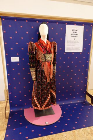 Ein ausgestelltes traditionelles Kleid auf einer Schneiderpuppe vor einem blauen Hintergrund mit rosa Mustern. Das Kleid hat einen bunten, auffälligen oberen Teil und einen langen Rock. Neben dem Kleid befindet sich eine Informationstafel.