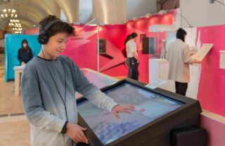 Ein junger Mann nutzt einen interaktiven Touchscreen in einem Ausstellungsraum. Er trägt Kopfhörer und konzentriert sich auf den Bildschirm. Im Hintergrund sind Besucher der Ausstellung zu sehen.