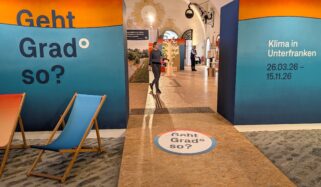 Eingang zur Ausstellung „Geht Grad so? Klima in Unterfarnken“. Auf dem Boden gibt es eine Leitspur für blinde und sehbehinderte Menschen. Eine Person betritt den Ausstellungsraum der hell, freundlich und interaktiv ist. Zwei Liegestühle stehen links.