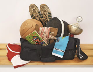 Eine Sporttasche mit verschiedenen Gegenständen, darunter ein Hallenfußball, ein Buch, ein Pokal, ein Netz und Turnschuhe auf einer Holzbank.