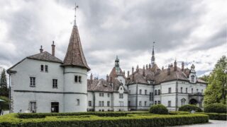 Eine historische Burg mit mehreren Türmen und Fachwerk in einer parkähnlichen Umgebung. Grüne Hecken umrahmen die Anlage, während der Himmel mit grauen Wolken bedeckt ist.