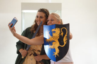 Zwei junge Frauen machen lachend ein Selfie von sich im Museum. Die vordere hält dabei ein buntes Ritterschild.